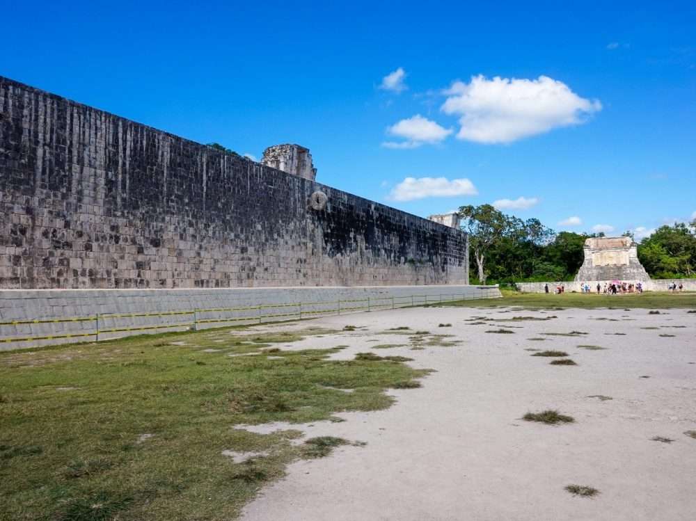 The Great Ballcourt at Chichén Itzá, featuring tall stone walls with embedded rings, surrounded by ancient ruins under a bright blue sky