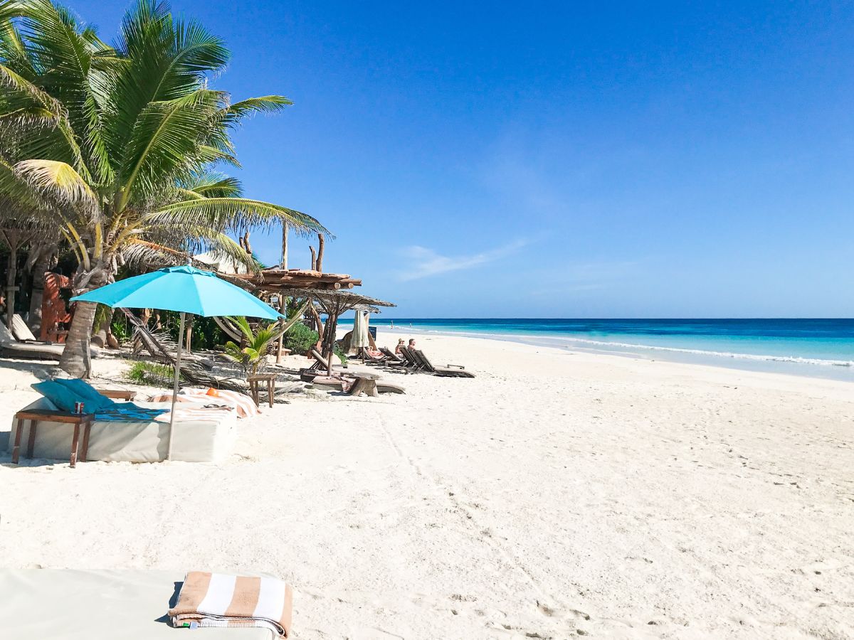 Idyllic beach in Tulum Mexico with white sand, a blue umbrella, lounge beds, palm trees, and a clear blue ocean under a sunny sky, perfect for a relaxing getaway.